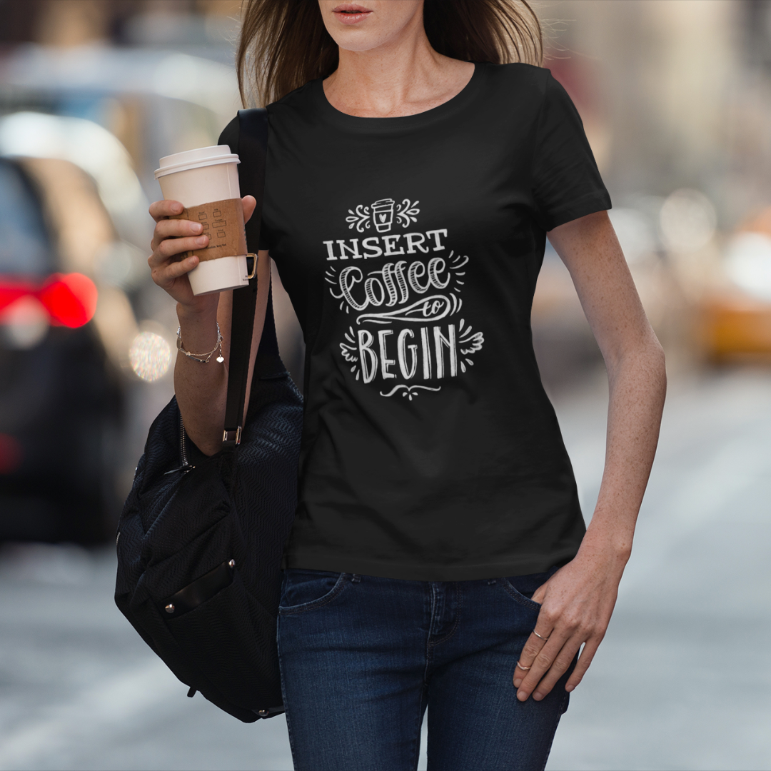 Woman walking down a busy street holding a coffee and wearing a t-shirt that says Insert Coffee to Begin