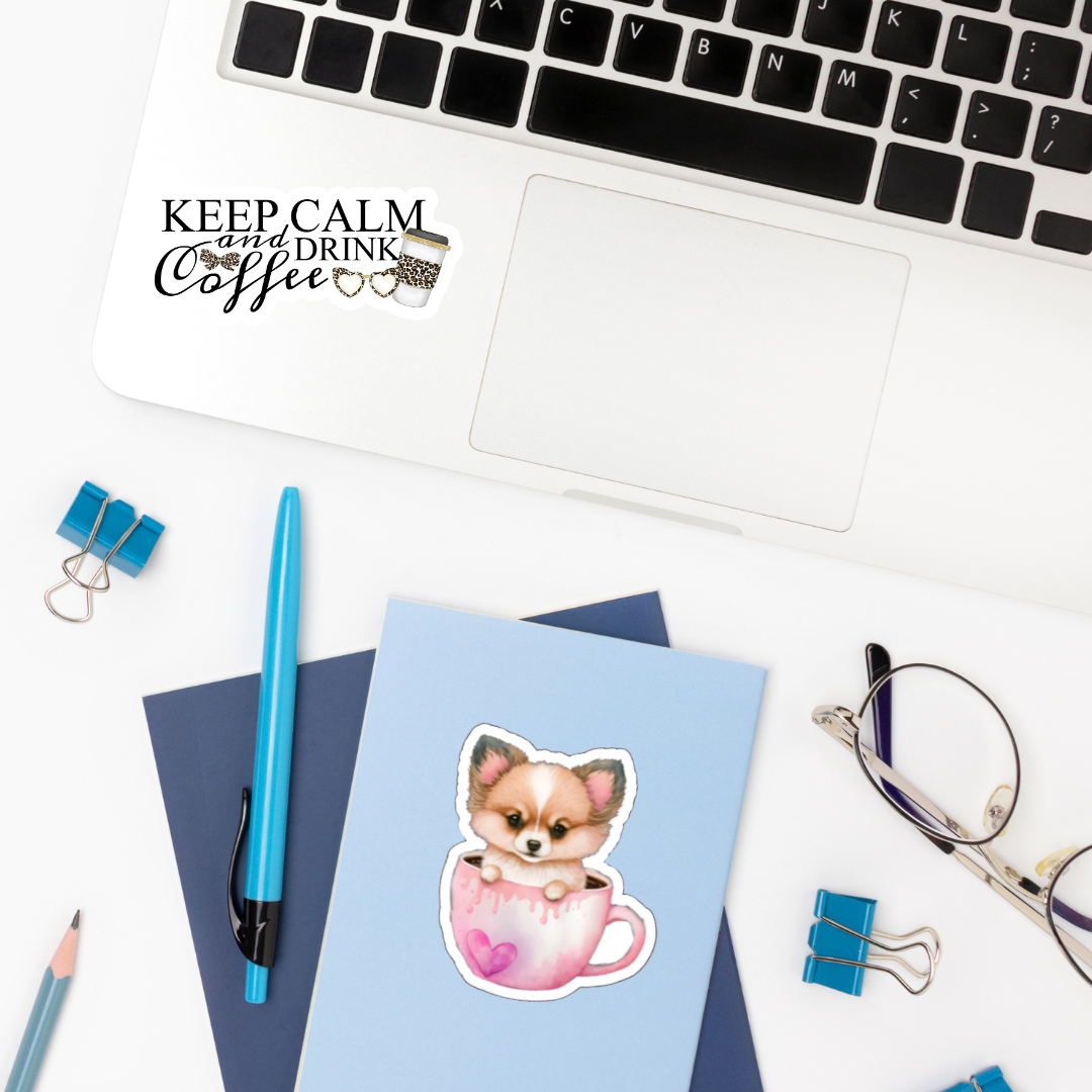 Desktop with various items on top with the focus on a sticker of a Corgi puppy sitting in a pink coffee mug done in watercolor.